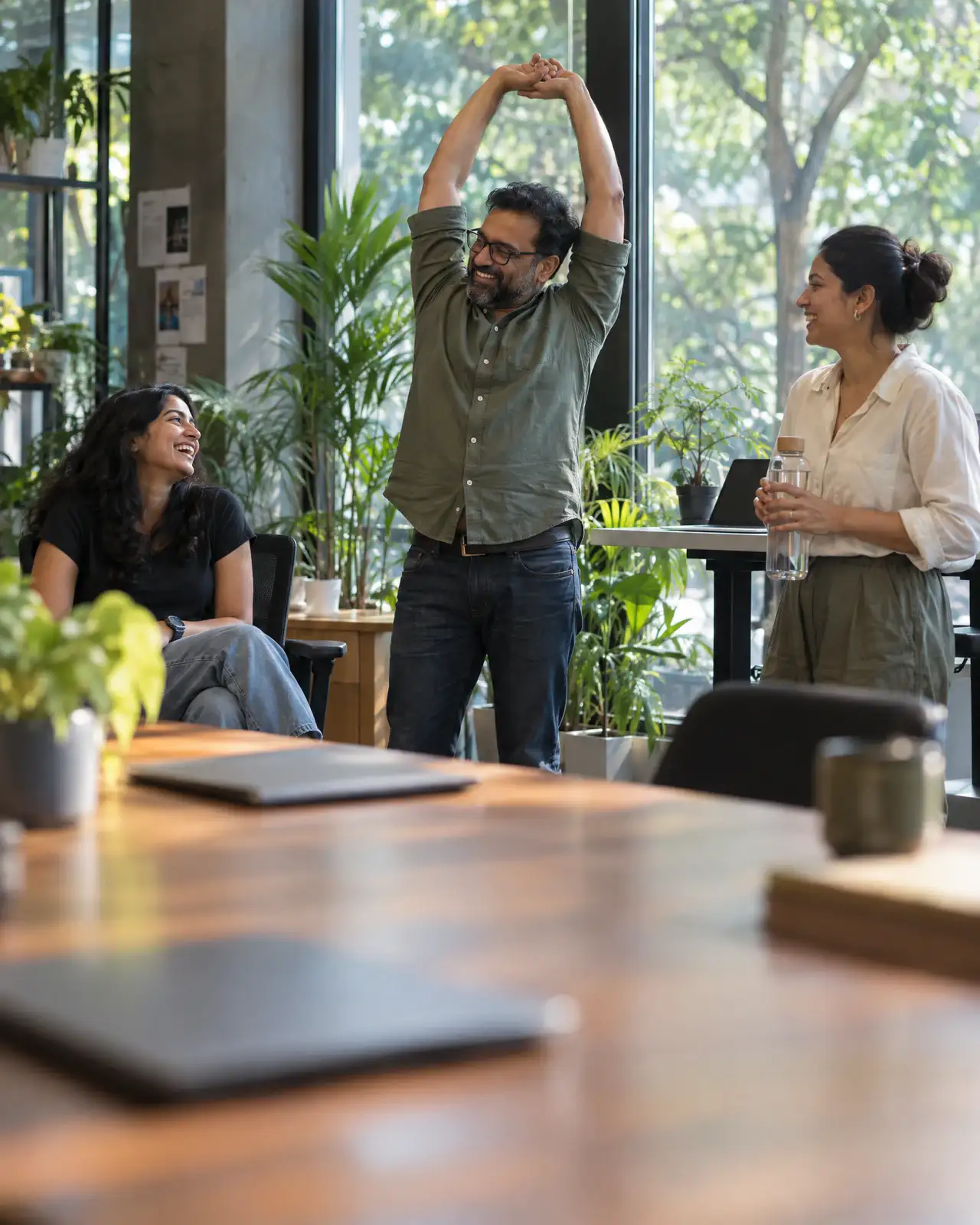 Colleagues laughing and stretching during a break in a bright office full of plants.